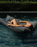 Person lying on a gray inflatable pillow in a pool with greenery in the background