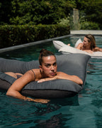 Two women relaxing on inflatable lounge chairs in a pool with greenery in the background